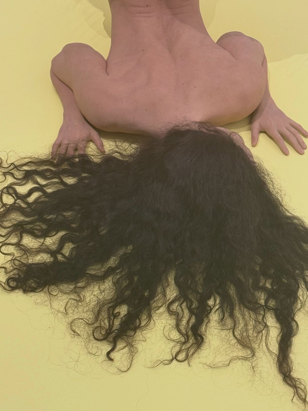노란 바닥에 엎드린 사람 모양 조각을 위에서 본 모습 — 맨 등과 바닥에 펼쳐진 길고 검은 곱슬머리